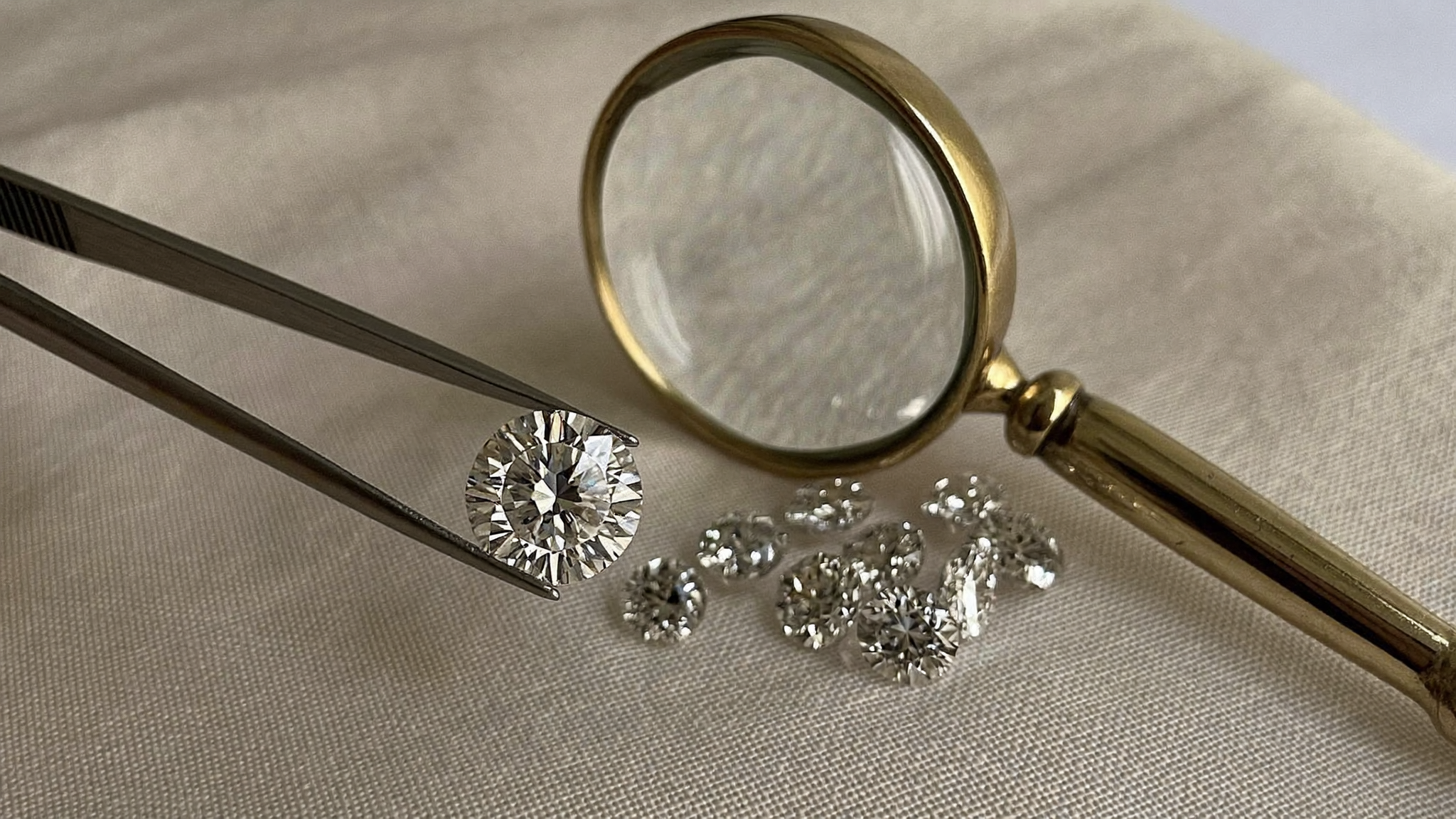 Lab grown diamond certification: jeweler's tweezers holding a lab-grown diamond beside a loupe on cream linen