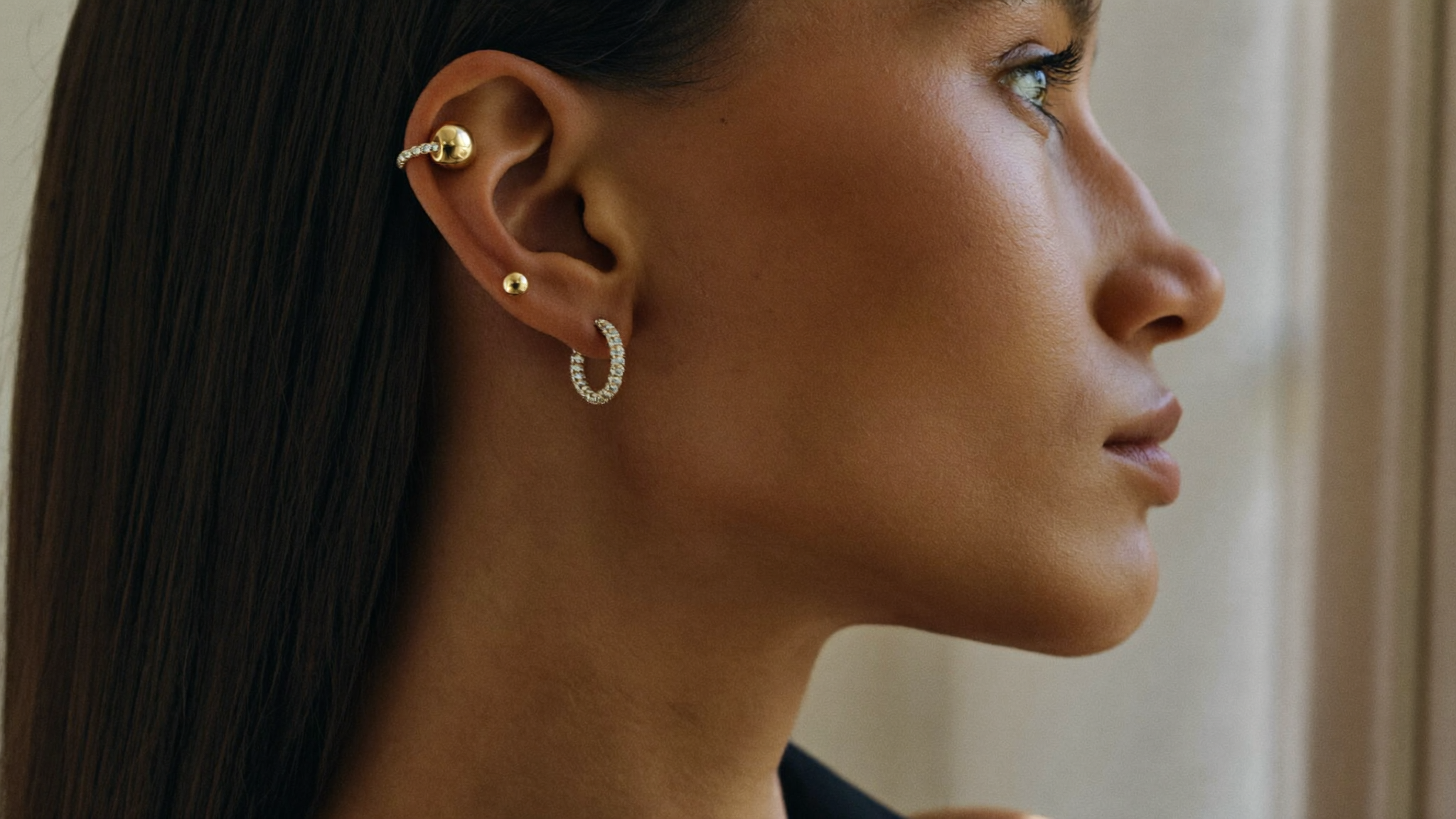 Curated ear stack of diamond stud, hoop, and ear cuff in solid gold, close-up in natural light