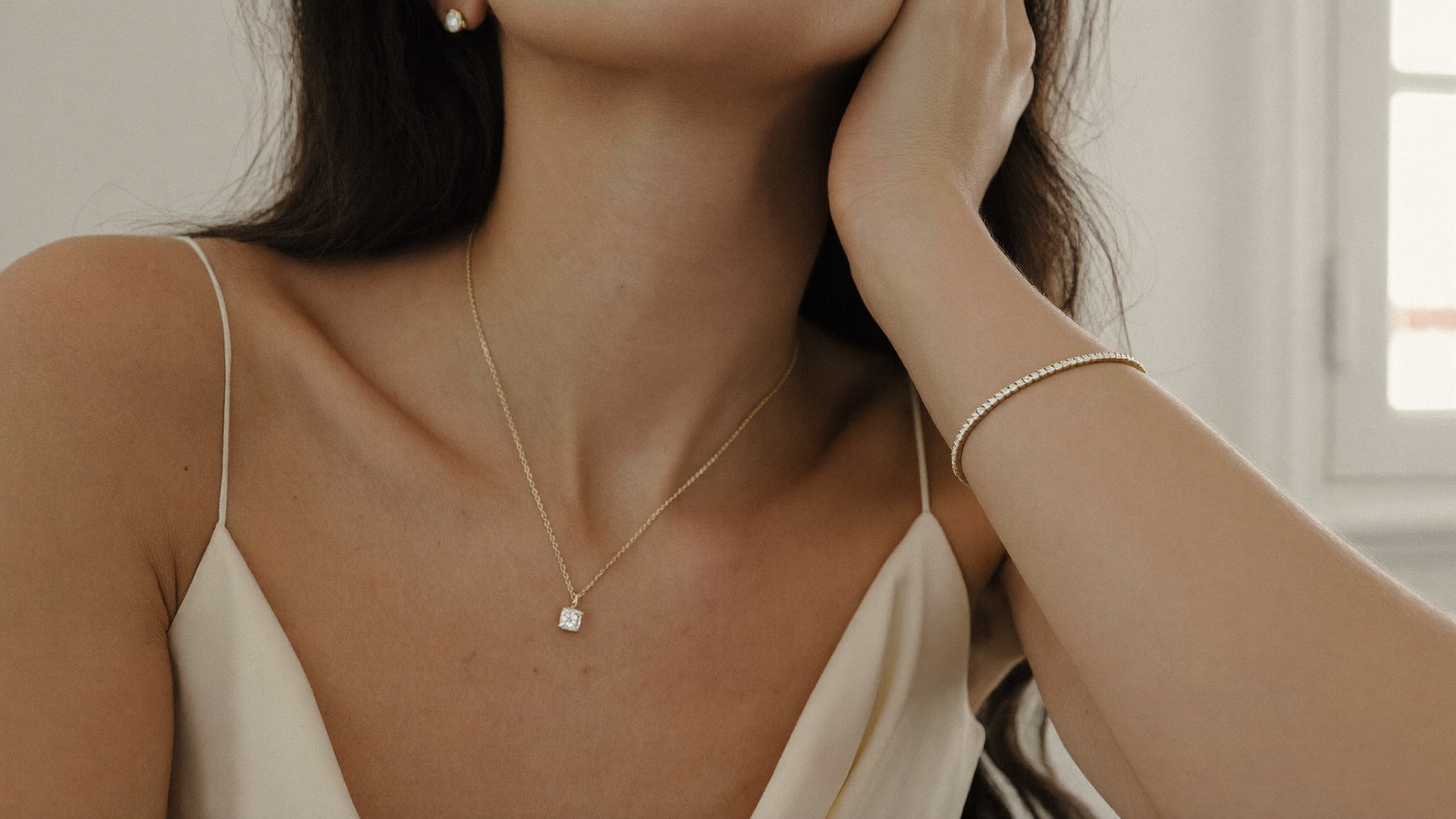 Woman in cream silk wearing a single diamond solitaire pendant, stud, and slim gold tennis bracelet, quiet luxury jewelry