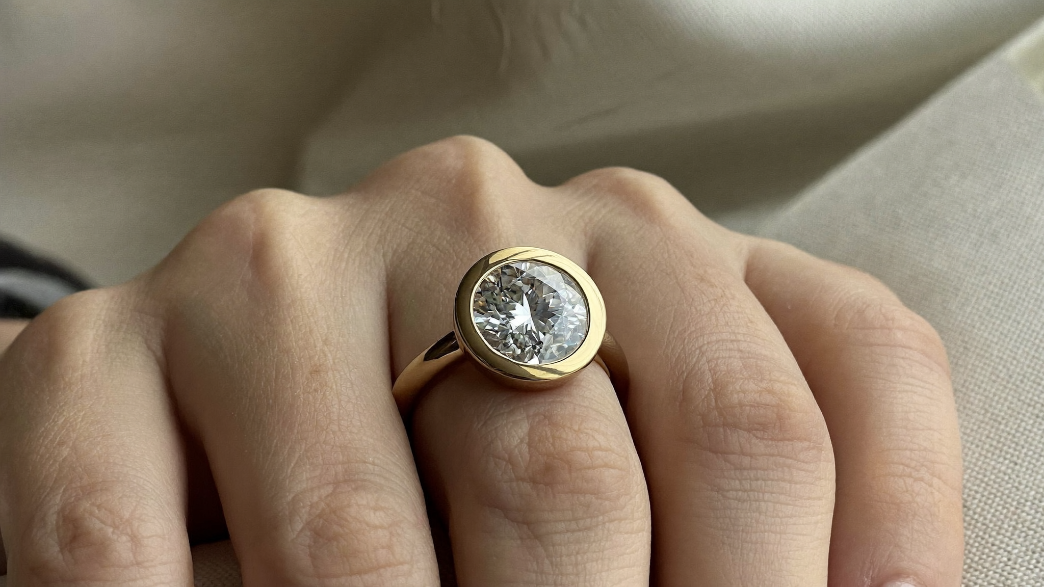 Close-up of a bezel set engagement ring in warm yellow gold on softly lit fingertips, natural light