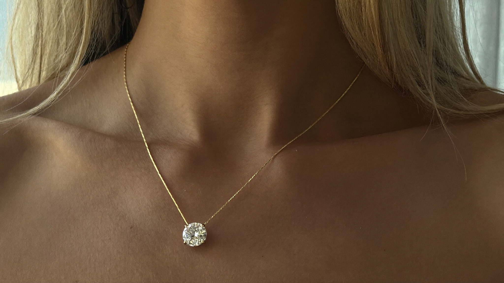 Close-up of a diamond solitaire necklace on a gold chain resting on a woman's collarbone in warm natural light